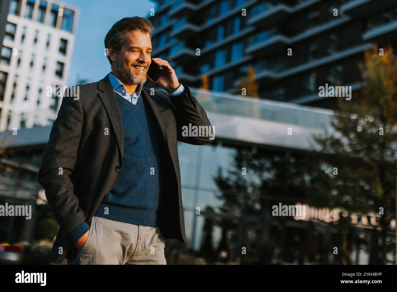 Ein gut gekleideter Mann schlendert selbstbewusst in einer urbanen Umgebung und führt ein lebhaftes Telefongespräch, während er an zeitgenössischer Architektur und vorbei geht Stockfoto