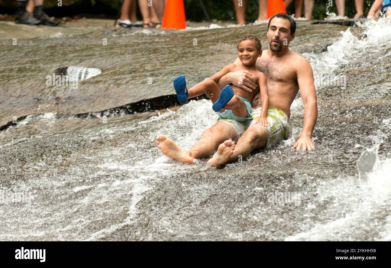 David und sein Sohn Preston Dougherty rutschen auf dem Sliding Rock im Pisgah National Forest, NC. Sliding Rock ist eine 60 Meter lange, rutschige Kaskade in den 50-60 Grad tiefen Pool. Es gibt 2 Aussichtsplattformen und Rettungsschwimmer im Dienst während der Saison. Dies ist ein stark genutztes Naherholungsgebiet, das sich oft voll ausfüllt. Personen, die am Straßenrand außerhalb des ausgewiesenen Parkplatzes parken, erhalten ein Ticket. Es wird auch empfohlen, alte Shorts und Turnschuhe zu tragen, wenn du dich für den Sprung entscheidest. Der Eintritt kostet 2,00 $ pro Person für Schieber und Beobachter. (Foto des Forstdienstes b Stockfoto