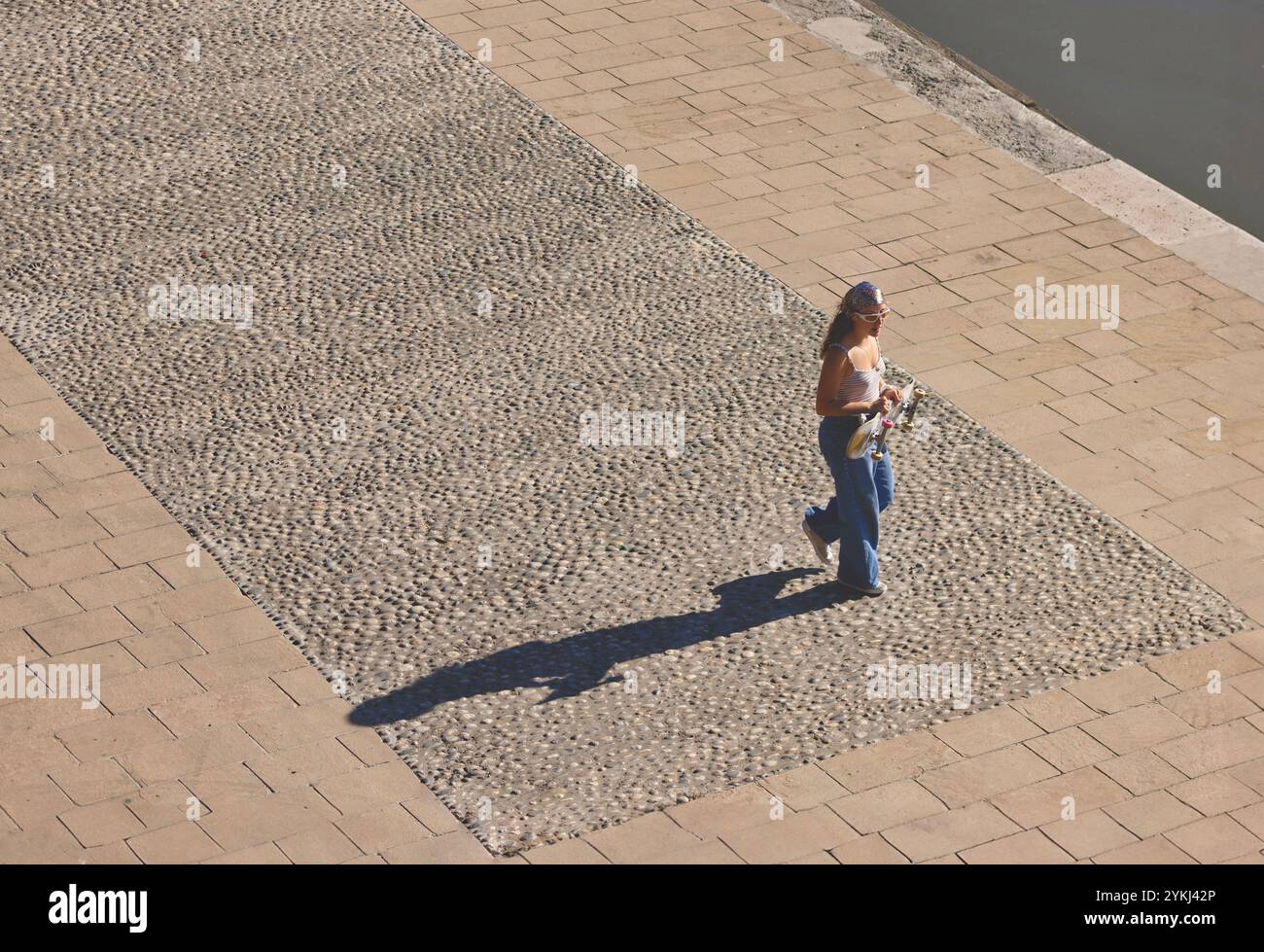 Eine Frau, die gemütlich auf einem bezaubernden Kopfsteinpflasterweg entlang eines wunderschönen Wasserkörpers spaziert Stockfoto