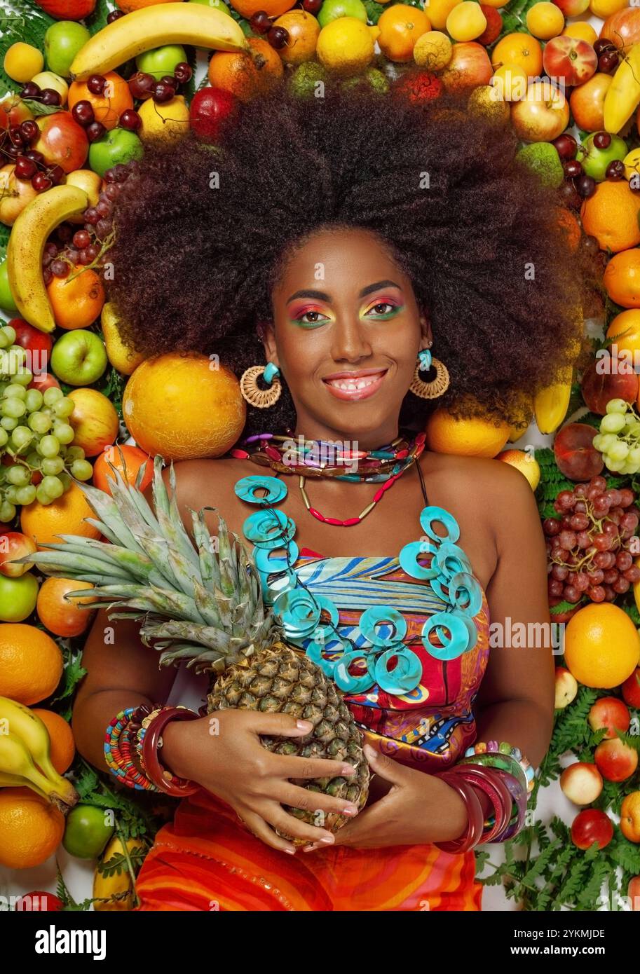 Die junge schöne afrikanische schwarze Frau lächelt in die Kamera, während sie im Studio zwischen Früchten liegt Stockfoto