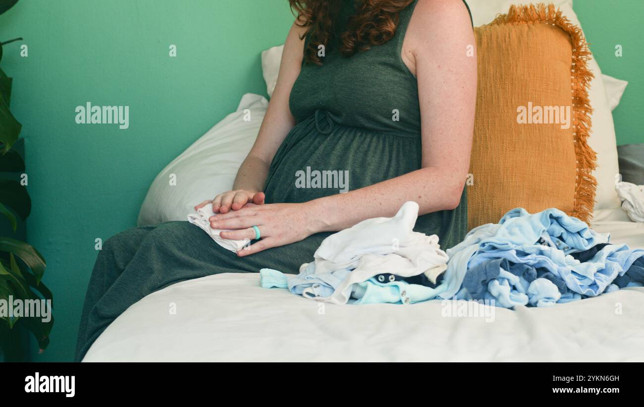 Schwangere Frau, die mit Babykleidung auf dem Bett sitzt, in friedlicher häuslicher Umgebung Stockfoto