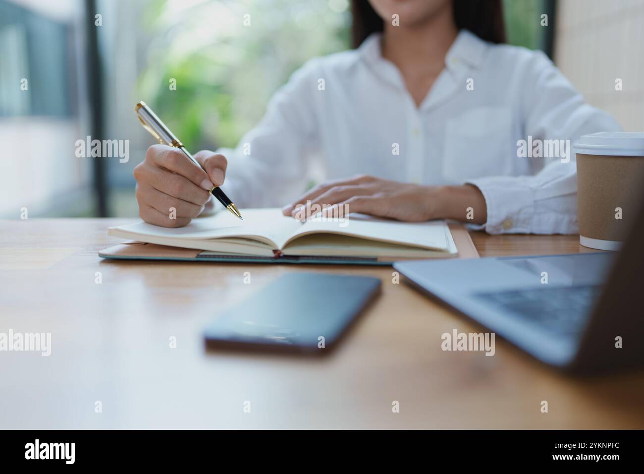 Professionelle Schreibnotizen in einem Notizbuch mit Stift, umgeben von einem Laptop und Kaffee, schaffen eine produktive Atmosphäre in einem hellen Arbeitsbereich. Stockfoto