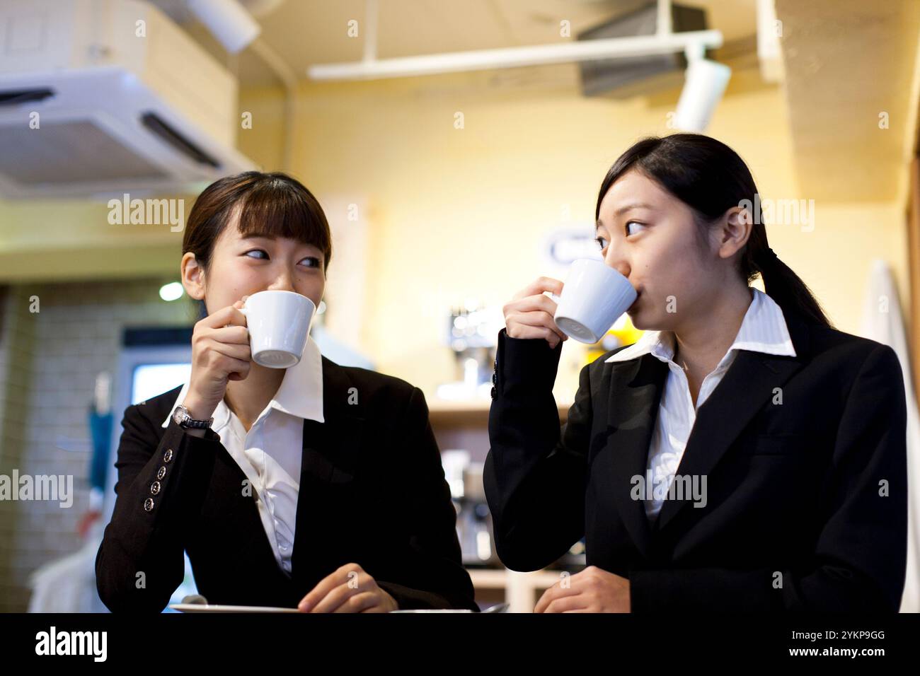 Zwei Frauen in Anzügen trinken Kaffee mit viel Milchschaum an einer Cafétheke. Stockfoto