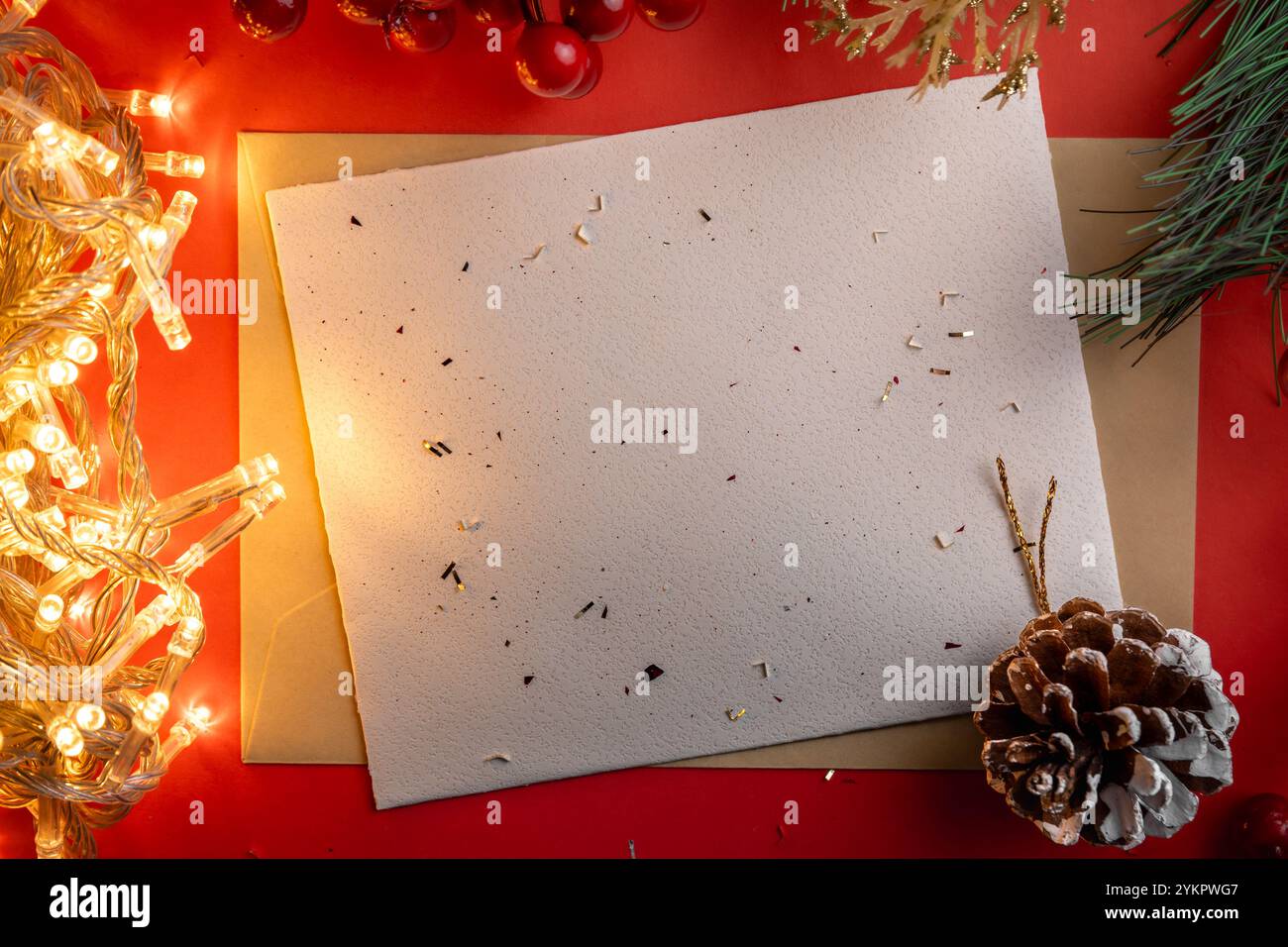 Eine leere Weihnachtskarte liegt auf einem roten Hintergrund, umgeben von festlichen Elementen wie einem Tannenzapfen, Tannenzweigen und warmen Lichterketten, die an eine gemütliche Atmosphäre erinnern Stockfoto