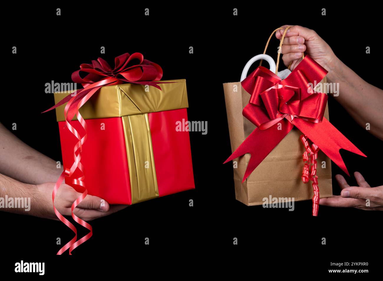 Nahaufnahme von zwei Händen, die eine rote Geschenkbox und eine Papiertüte mit roten Bändern auf schwarzem Hintergrund austauschen, die Weihnachtsstimmung symbolisieren. Stockfoto