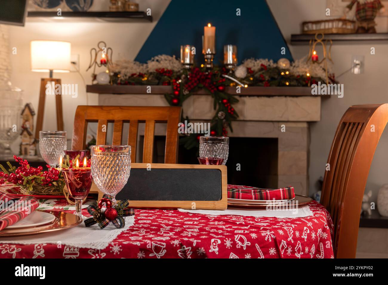 Charmante Feiertagsspeisenanlage mit roter Tischdecke, elegantem Glas und festlicher Einrichtung durch einen warm beleuchteten Kamin, die eine gemütliche Umgebung schafft. Stockfoto