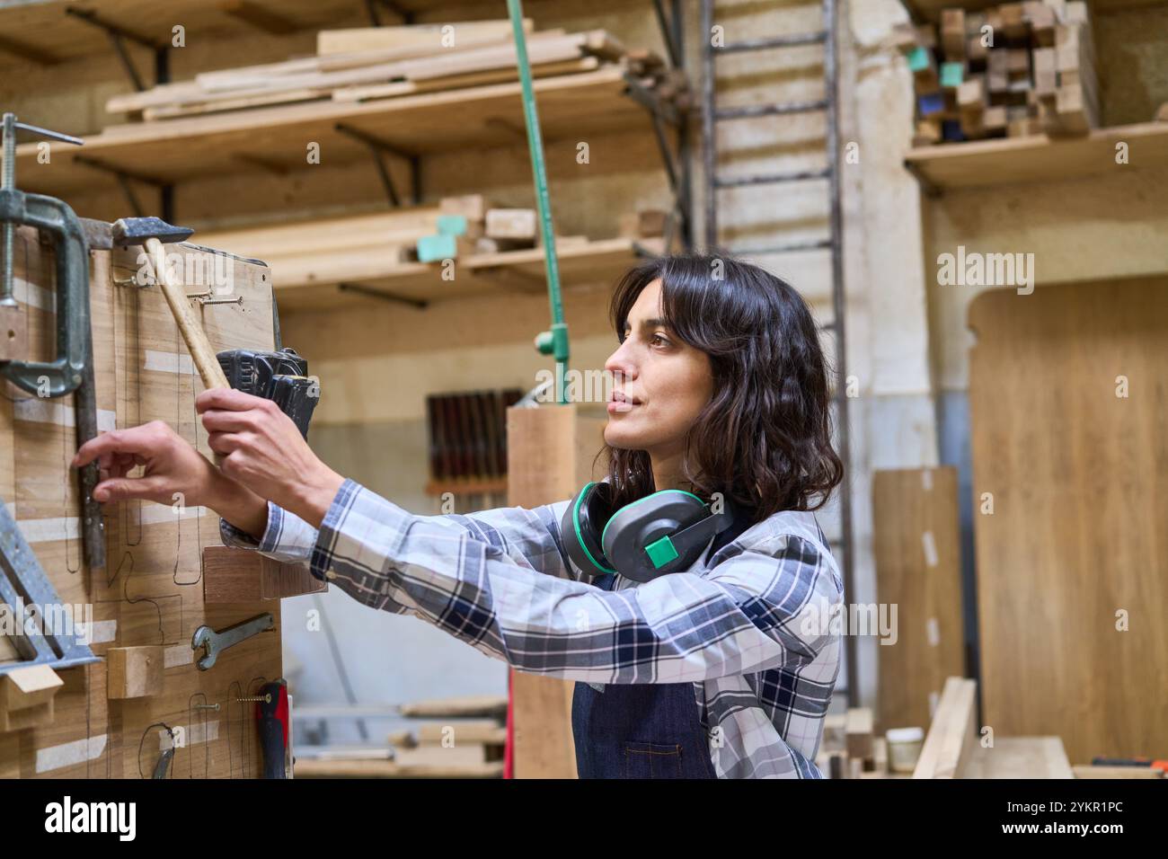 Eine junge Frau in einem Holzhof arbeitet aufmerksam mit Werkzeugen und Holz und zeigt sorgfältige Handwerkskunst. Ihr fokussierter Ausdruck unterstreicht ihre Stockfoto