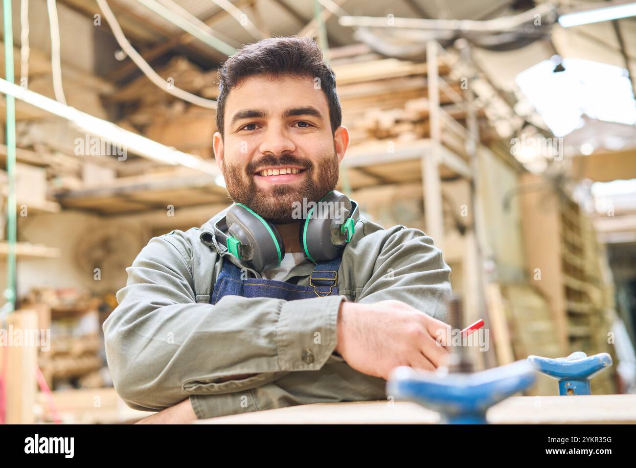 Ein junger Mann steht selbstbewusst in einem belebten Holzhof, trägt Kopfhörer und Arbeitskleidung. Sein Lächeln spiegelt den Geist der Teamarbeit und der Entwicklung von Fähigkeiten wider Stockfoto