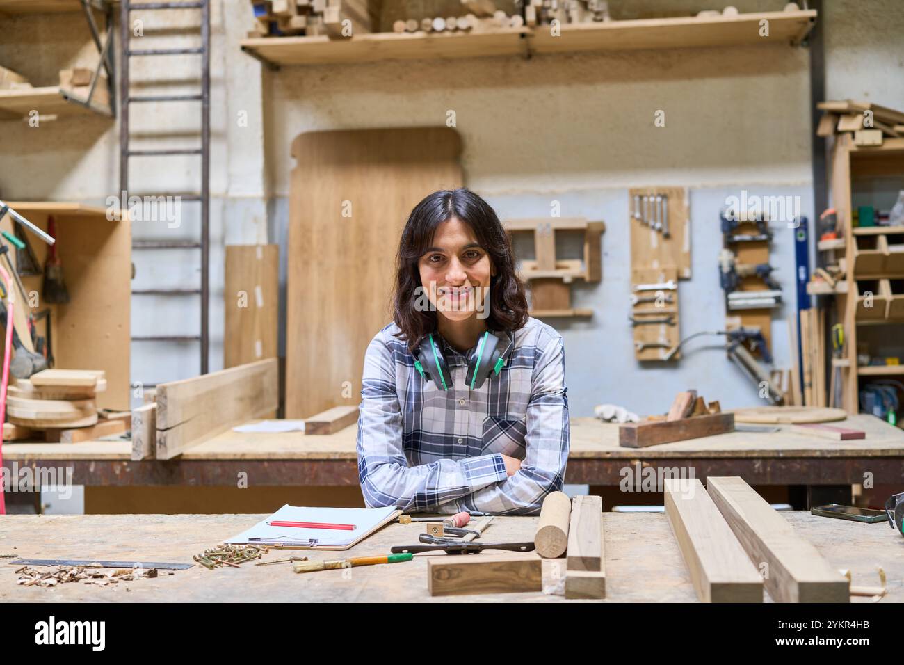 Ein junger Zimmermann lächelt zuversichtlich, während er in einer geschäftigen Werkstatt von Holzmaterialien umgeben ist. Das Bild fängt das Wesen der Handwerkskunst ein, Hingabe Stockfoto