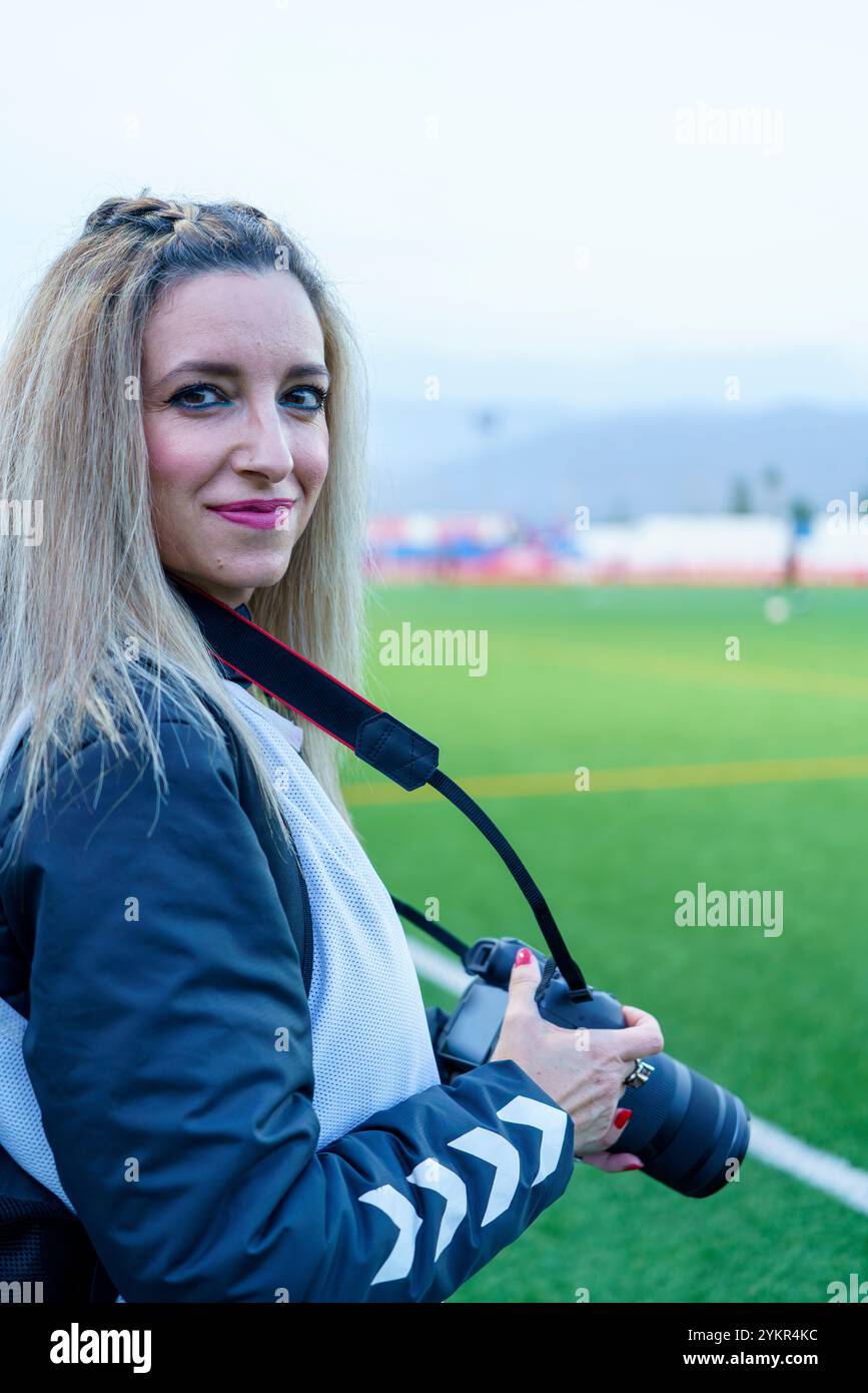 Sportfotografin mittleren Alters lächelt die Kamera an, während sie auf einem Fußballfeld in der Dämmerung Ausrüstung hält Stockfoto