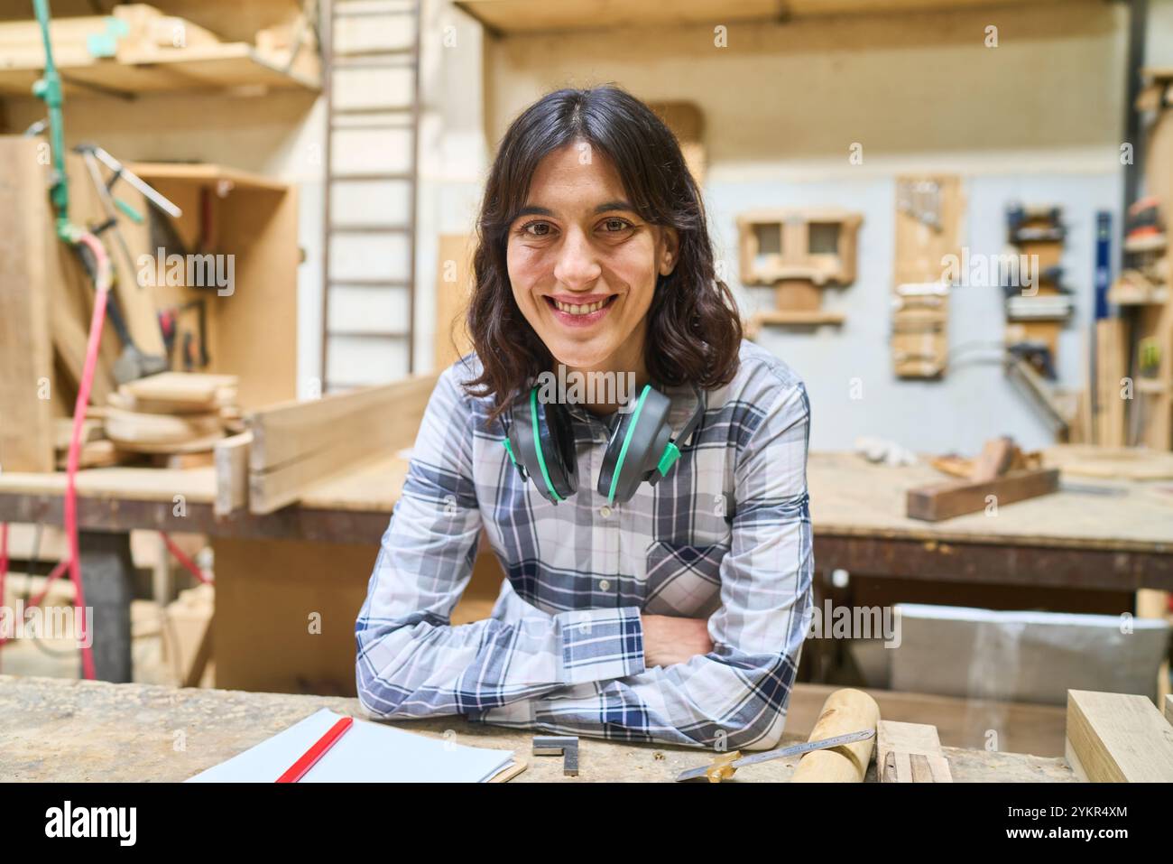 Eine junge Frau lächelt selbstbewusst in einer Holzwerkstatt, umgeben von Holzwerkzeugen. Ihr entspanntes Auftreten deutet auf Fachwissen und Begeisterung hin Stockfoto