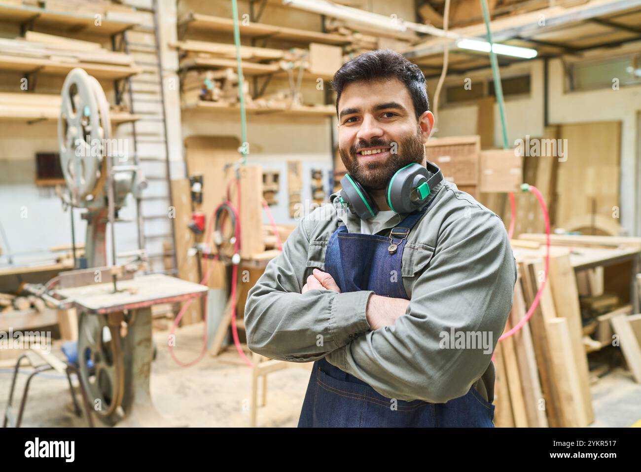 Junger Zimmermann, der stolz in einer Holzwerkstatt steht, umgeben von Werkzeugen und Holz, zeigt Vertrauen und Handwerkskunst in einem professionellen lumb Stockfoto