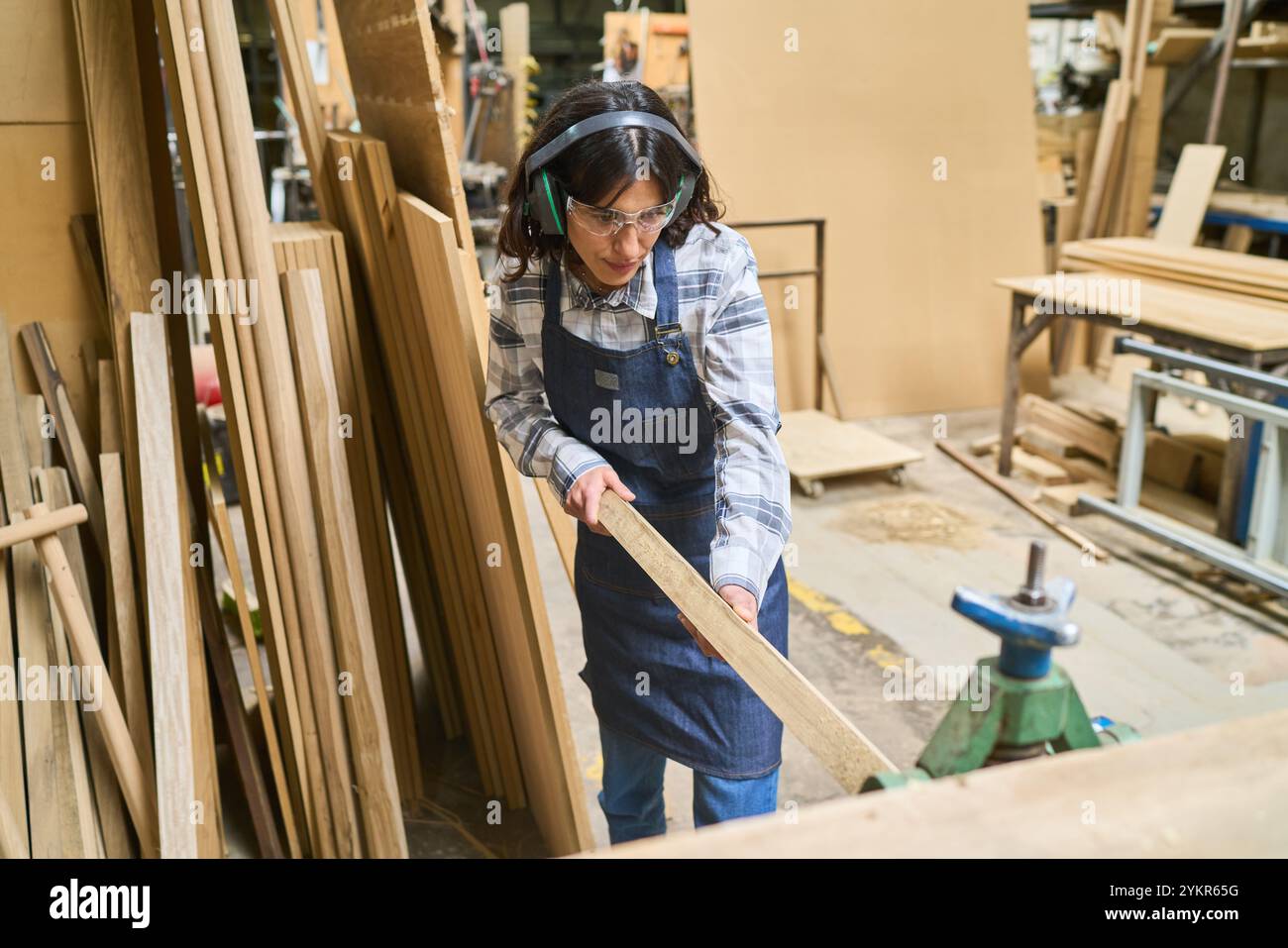 Ein junger Arbeiter konzentriert sich auf den Umgang mit Holz in einem Holzhof und demonstriert Handwerkskunst und Sicherheit mit Schutzbrillen und Kopfhörern. Diese Szene emp Stockfoto