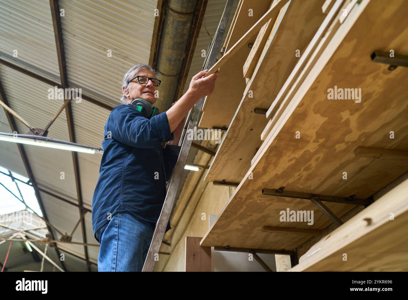 Ein erfahrener Arbeiter, der Sicherheitsausrüstung trägt, organisiert Holzdielen in einem belebten Holzhof. Betont die Bedeutung von Sicherheit und Organisation in Arbeitsaufträgen Stockfoto