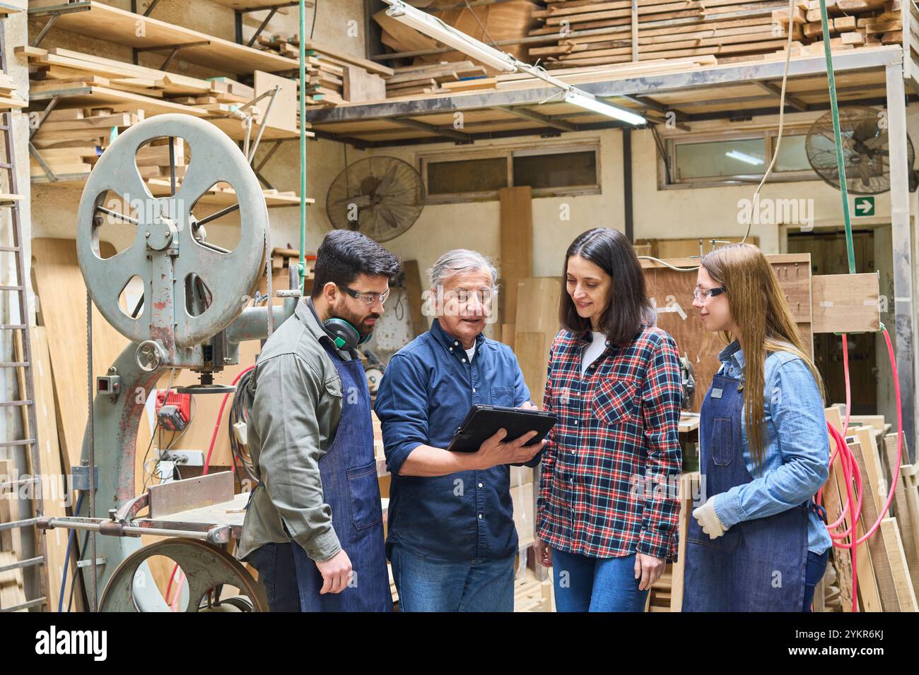 Jüngere und ältere Arbeitnehmer arbeiten in einem Holzlager zusammen und konzentrieren sich dabei auf Schulungen und Teamarbeit. Das geschäftige Werkstattumfeld hebt die Ausbildung und hervor Stockfoto