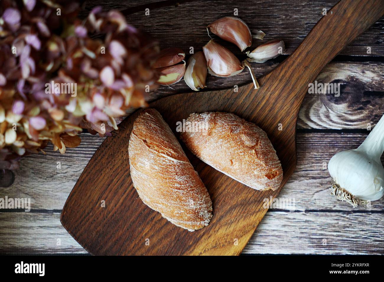 Rustikale Brötchen auf einem Holzbrett mit Knoblauchzehen und getrockneten Blumen im Hintergrund. Gemütliche Komposition im Bauernhausstil. Stockfoto