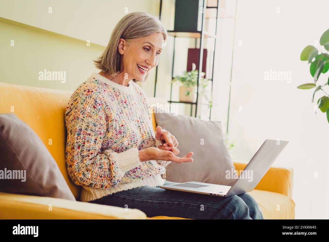 Glückliche ältere Frau, die zu Hause einen entspannenden Videoanruf genießt, in einem gemütlichen Wohnzimmer mit natürlichem Tageslicht und charmanter Inneneinrichtung Stockfoto