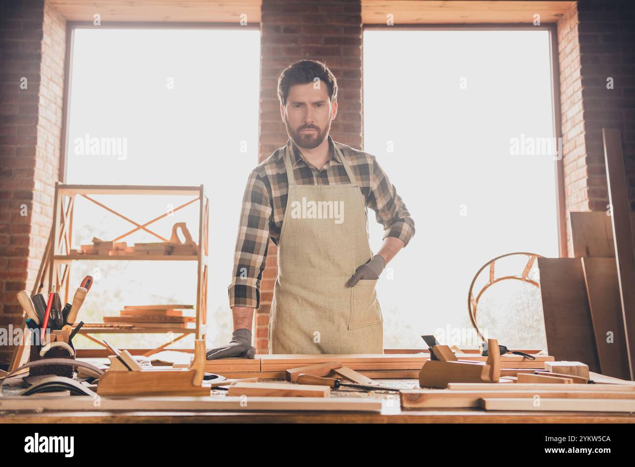 Foto eines starken, selbstbewussten bärtigen Mannes mit spezieller Schürze und Handschuhen, der in der Nähe seiner Handwerker oder hölzernen Flugzeugmodelle posiert Stockfoto