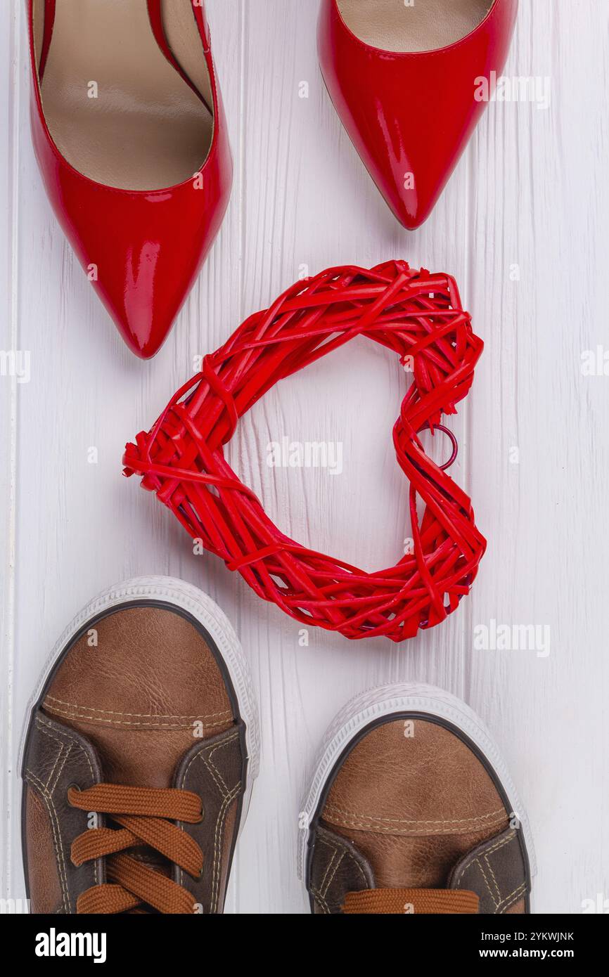 Vertikale Aufnahme von Draufsicht Schuhe und Herz. Rote Damenschuhe und braune Herrenschuhe auf weißem Schreibtisch Stockfoto