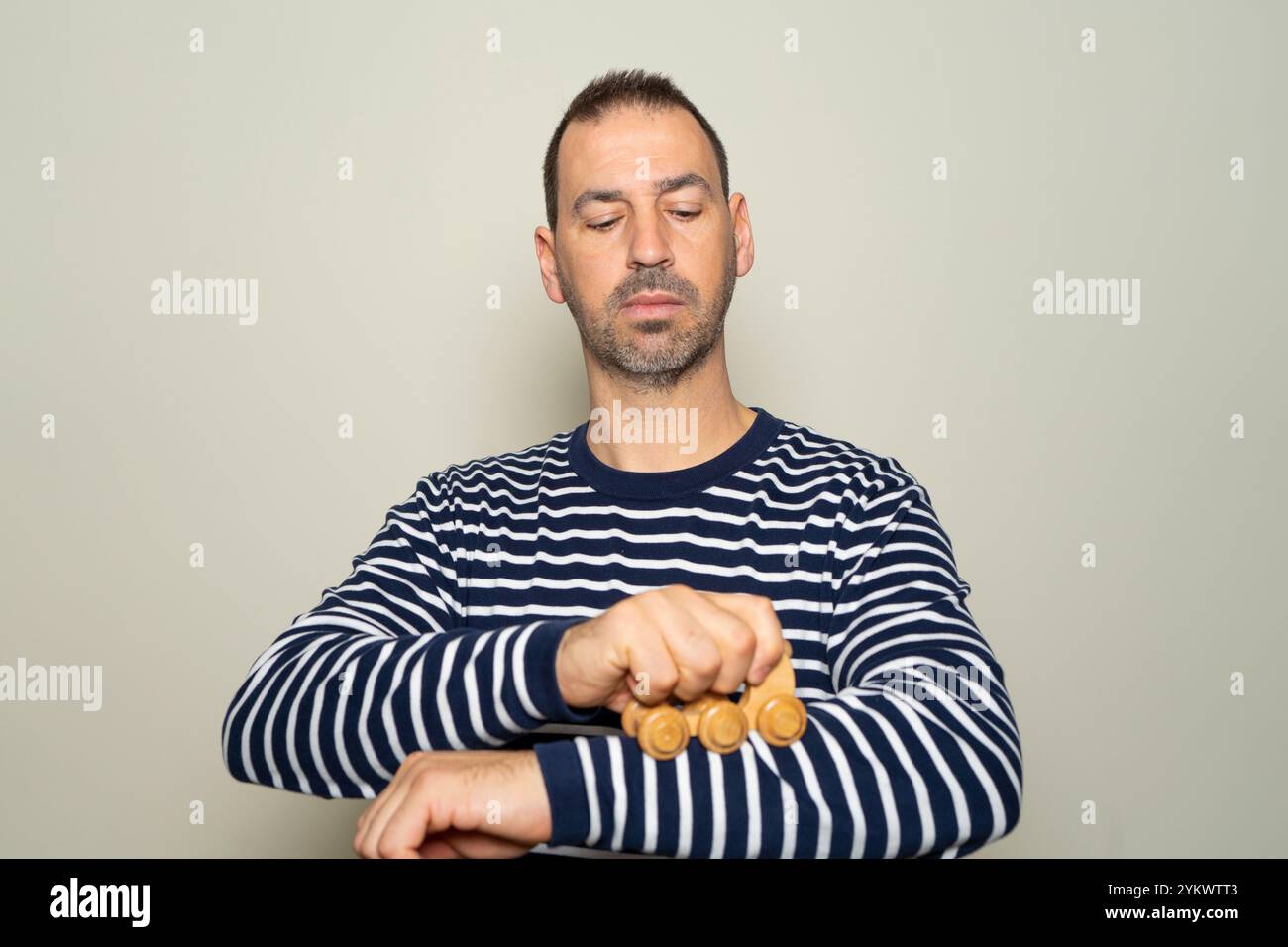 Bärtiger hispanischer Mann in seinen 40ern, der einen gestreiften Pullover trug, der ein hölzernes Massagegerät über seinen schmerzenden Arm führte, isoliert auf beigefarbenem Studiohintergrund Stockfoto