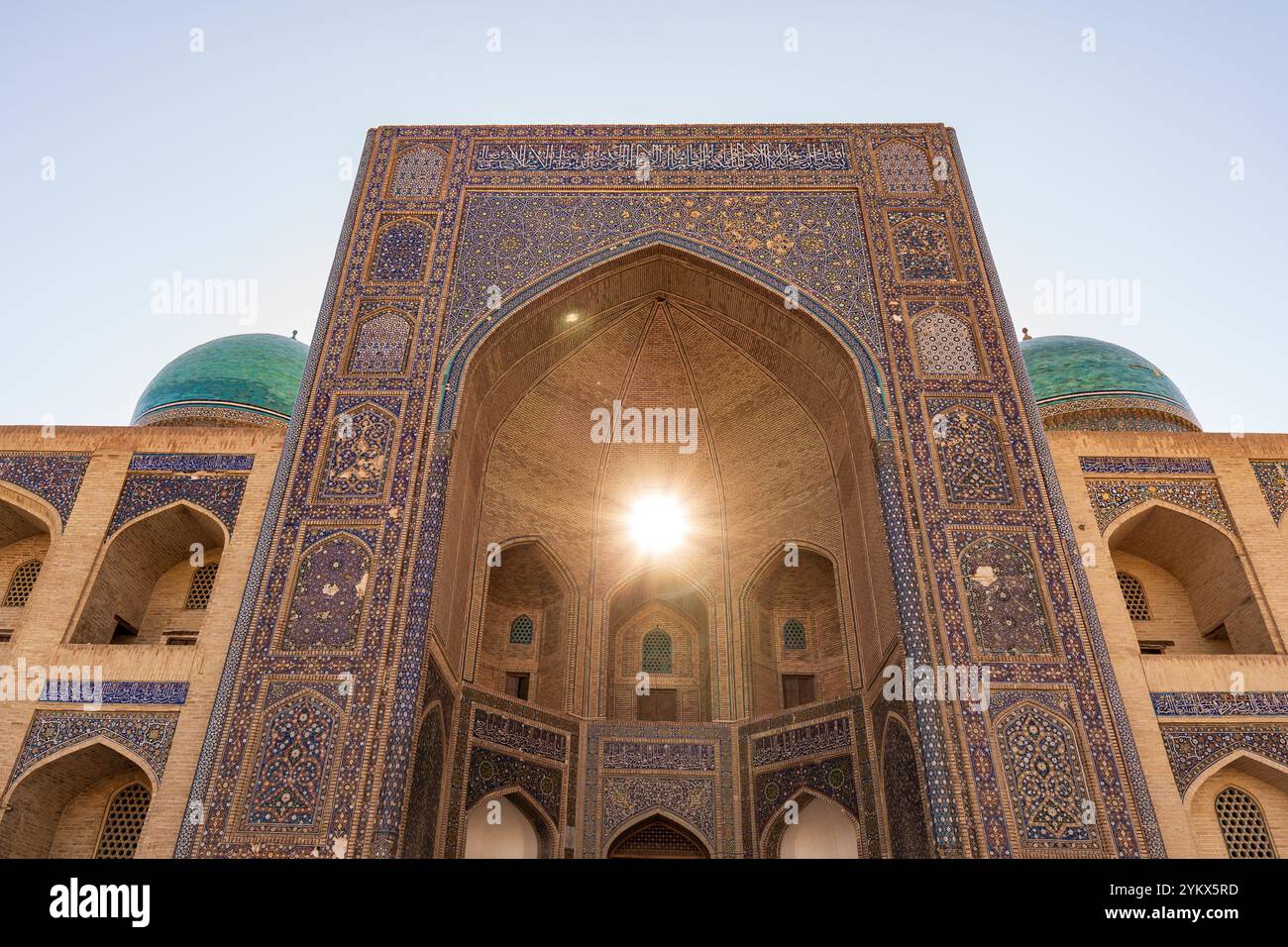 Goldenes Sonnenlicht strömt durch den zentralen Bogen der mir-i-Arab Madrasa, eingerahmt von den berühmten grünen Kuppeln in Buchara Stockfoto