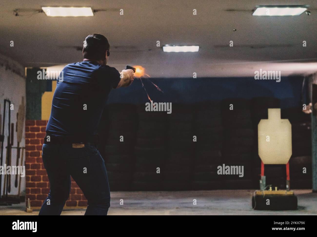 Eine Person, die eine Pistole auf einen Schießstand im Innenbereich feuert, mit humanoidem Ziel und sichtbarem Mündungsblitz. Ziel und Feuer, Training. Schusswaffe Stockfoto