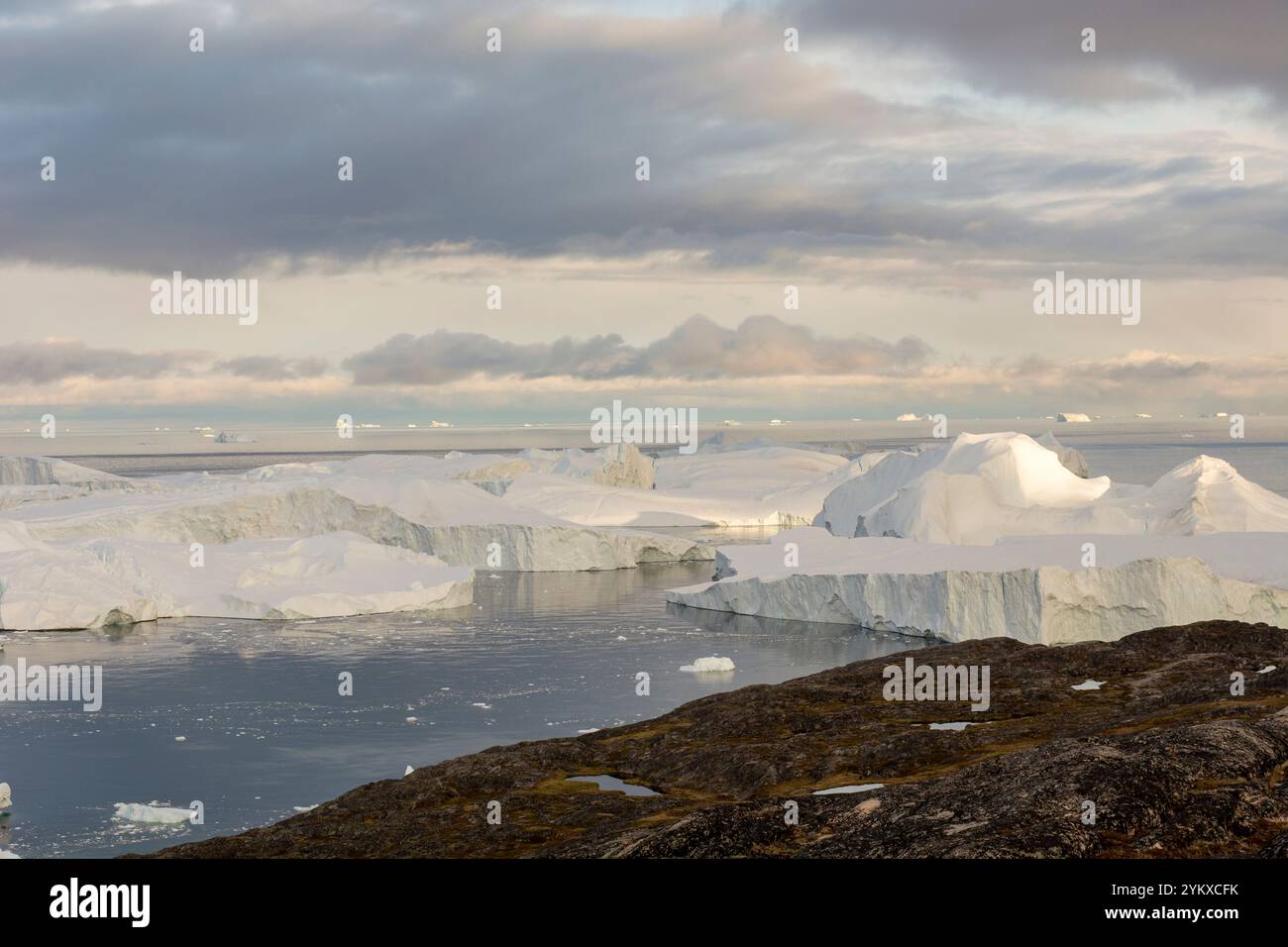 Die atemberaubende Landschaft von Ilulissat, Grönland, entfaltet sich mit einer dramatischen Mischung aus hoch aufragenden Eisbergen und zerklüfteten Felsformationen. Im Vordergrund, gezackt Stockfoto