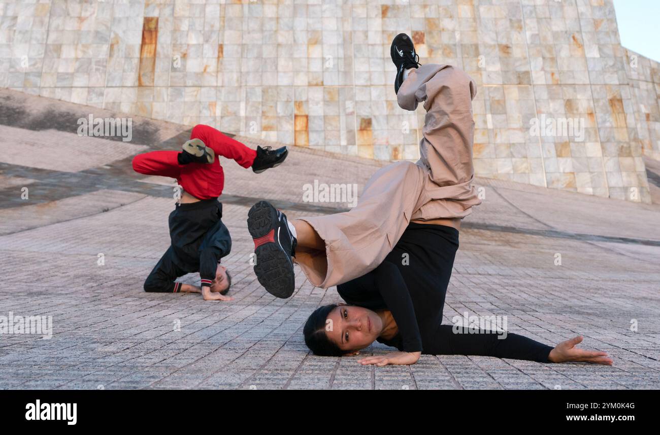 Eine Frau in einem schwarzen T-Shirt und einer braunen Hose, die eine Breakdance-Pose auf dem Boden macht. Ein Mann in roter Hose und schwarzem Sweatshirt schlägt eine andere Pose hinter sich Stockfoto