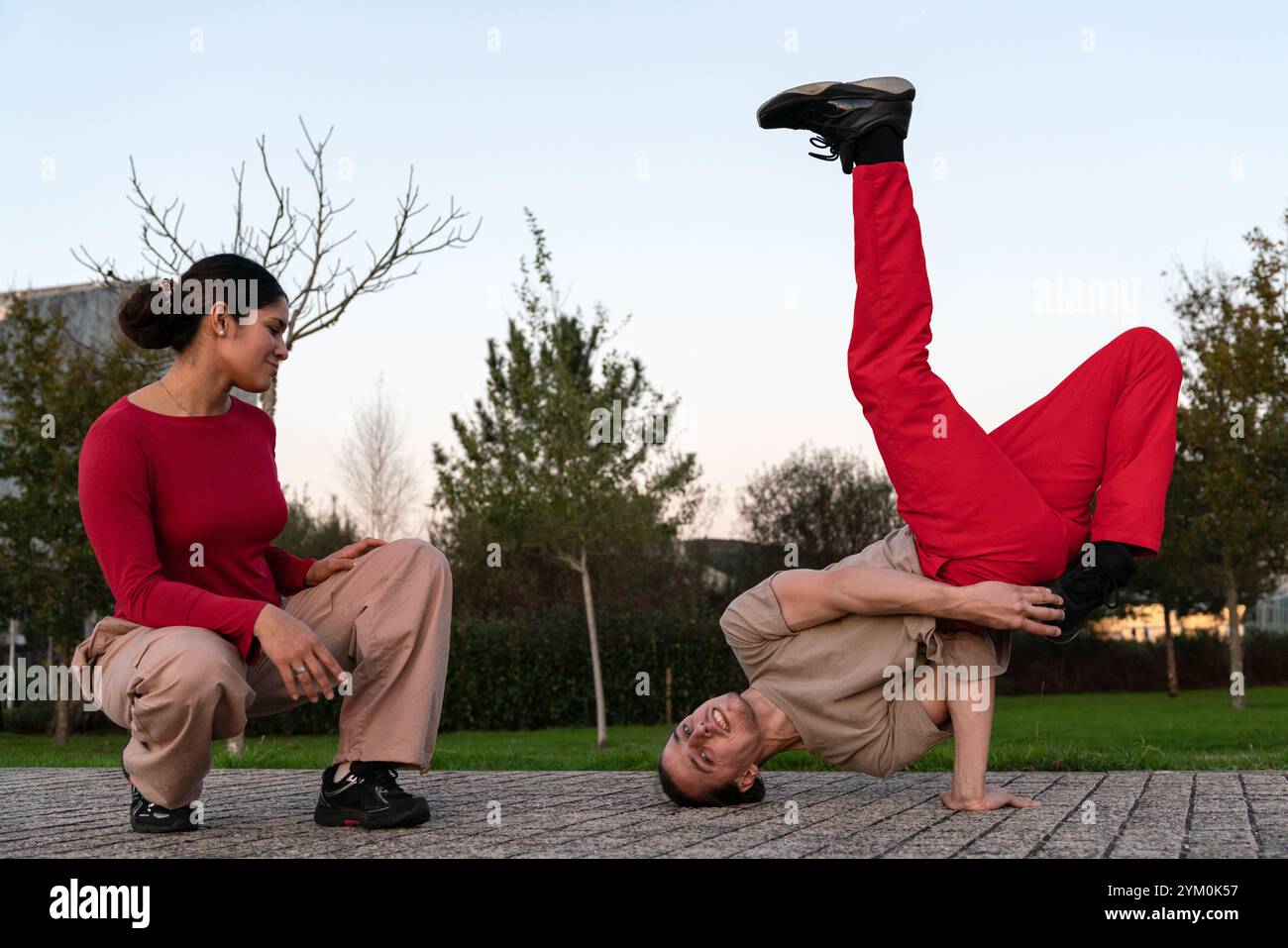 Ein Mann in roter Hose in einer Breakdance-Pose im Park, während eine Frau ihn beobachtet. Die Szene ist lässig und entspannt, Mann und Frau lächeln und engagieren sich Stockfoto