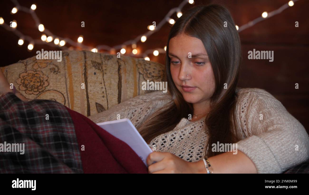 Teenager-Mädchen, das ein Buch liest, sitzt zu Hause auf der Couch bei schwacher Beleuchtung mit Weihnachtsbeleuchtung im Hintergrund Stockfoto