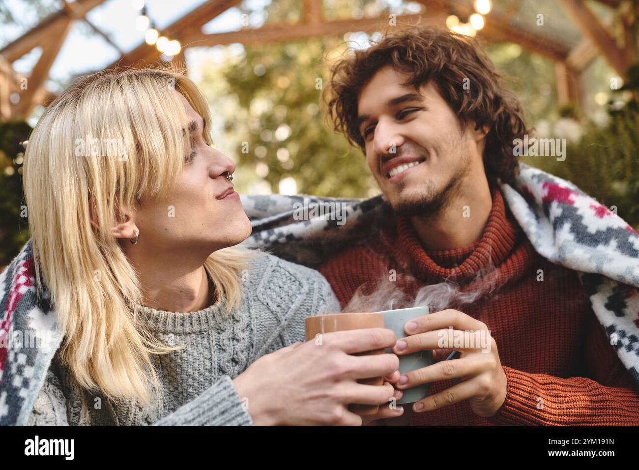 In einer ruhigen Umgebung im Freien genießen sich zwei junge und gutaussehende Männer in Gesellschaft, während sie warme Getränke teilen. Die gemütliche Atmosphäre, verstärkt durch ein blanke Stockfoto