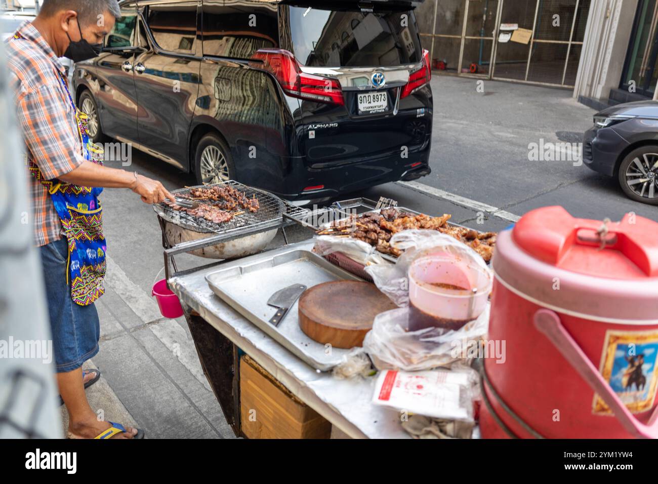 Ein Straßenverkäufer in Bangkok, Thailand, grillt Fleisch auf einem einfachen Grillstand am Straßenrand. Der Mann trägt eine karierte Hemd-Schürzen-Kombination und arbeitet konzentriert. Im Vordergrund sind Zutaten, ein Holzschneidebrett und eine rote Thermoskanne zu sehen, während im Hintergrund Autos und urbane Architektur die typische Straßenatmosphäre der Stadt einfangen. Dieses Foto spiegelt die authentische Esskultur und das geschäftige Straßenleben Bangkoks wider. *** Ein Straßenverkäufer in Bangkok, Thailand, grillt Fleisch an einem einfachen Barbecue-Stand am Straßenrand der Mann trägt ein kariertes Hemd und ap Stockfoto