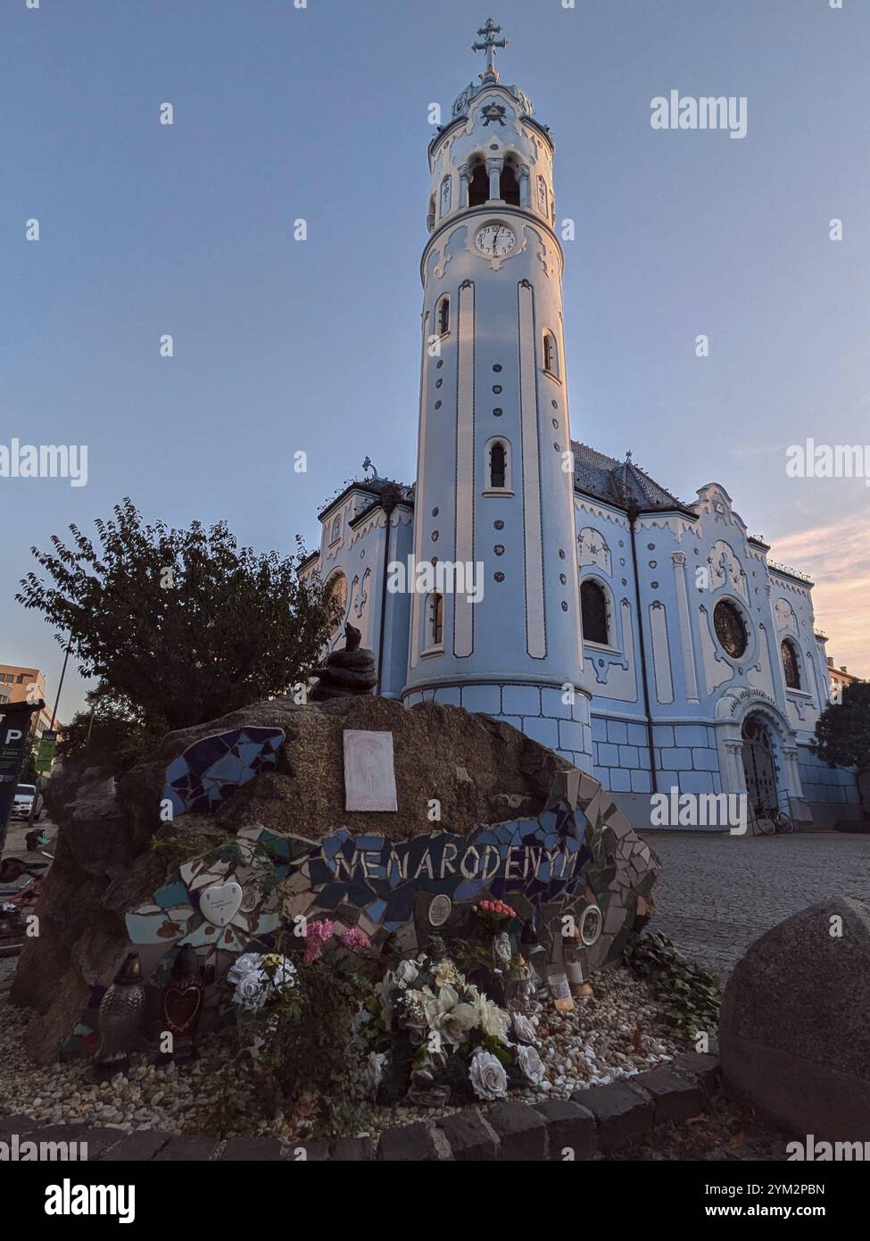 Eine blaue Kirche mit einzigartiger Architektur und ein Denkmal mit Blumen in der Dämmerung. Bratislava, Slowakei, Europa Stockfoto