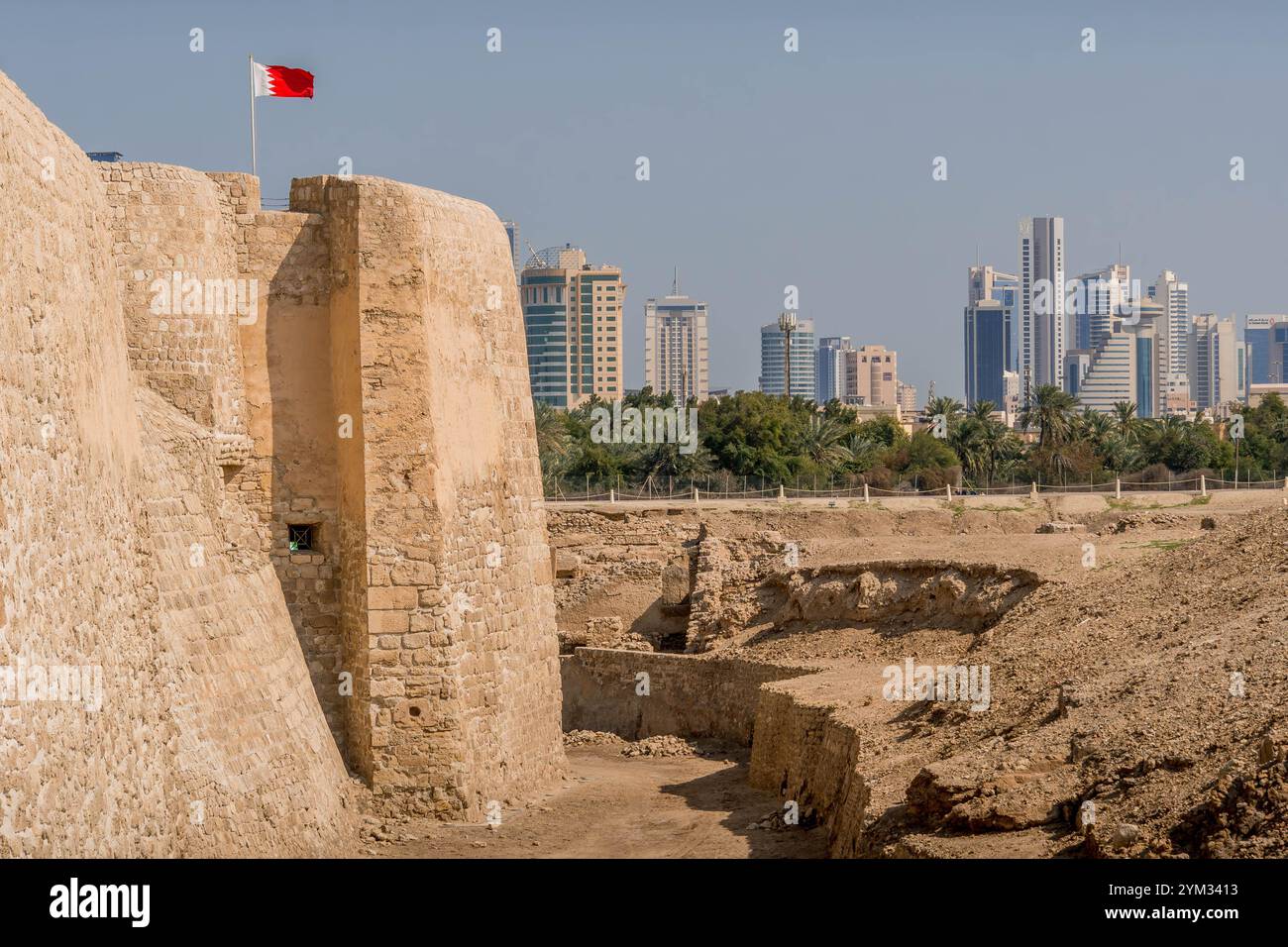 Die Mauer von Qal'at al-Bahrain (Fort Bahrain), eine archäologische Stätte in Manama, UNESCO-Weltkulturerbe mit der Skyline der Stadt und der Flagge Bahrains. Stockfoto