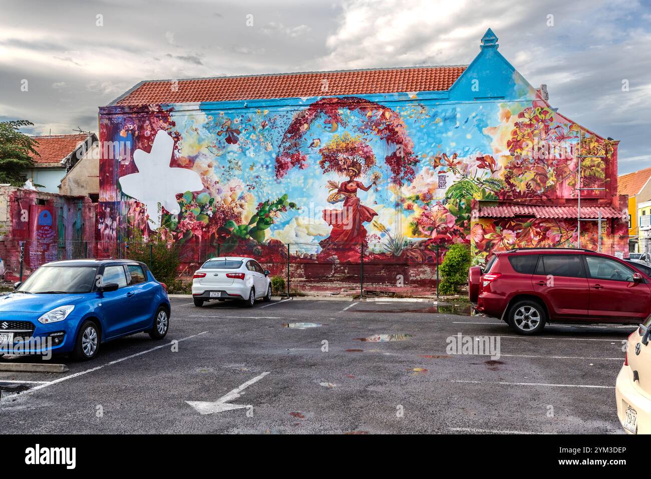 Das Wandgemälde „Djosa“ von Francis Sling bedeckt die Fassade eines Hauses im Otrabanda-Bezirk von Willemstad, Curaçao. Es wurde 2024 für das KayaKaya Murals Festival geschaffen. Das großflächige Werk zeigt eine Frau in einem roten Kleid inmitten tropischer Blumen – Bougainvillea, Kakteen, Hibiskus – und ein weißes Kreuz. Der Titel, in Papiamentu, erforscht die Frage nach Gott und Geschlecht. Stockfoto