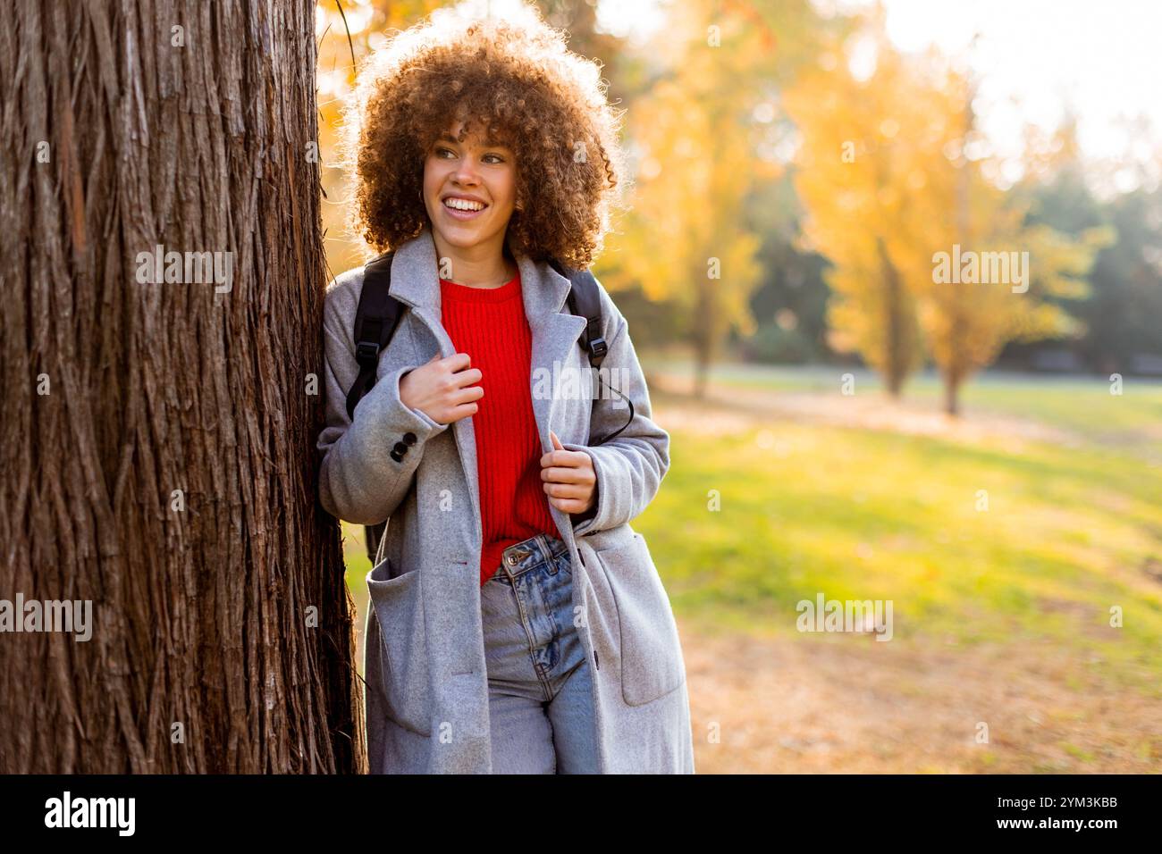 Eine lächelnde junge Frau mit lockigem Haar steht vor einem Baum und trägt ein gemütliches Outfit. Sie liebt die Schönheit des Herbstes in einem ruhigen Park voller Colo Stockfoto