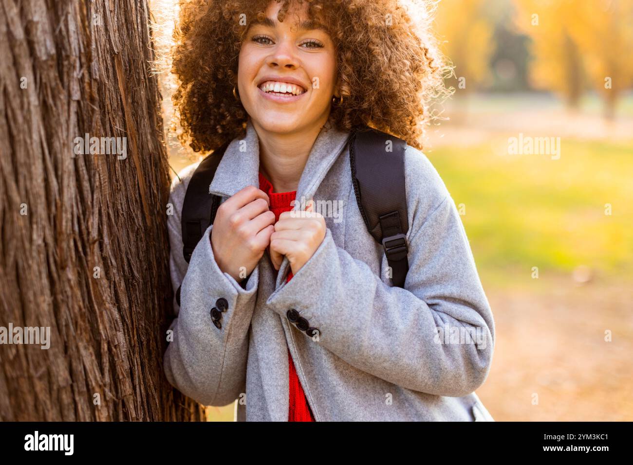 Eine lächelnde junge Frau mit lockigem Haar steht vor einem Baum und trägt ein gemütliches Outfit. Sie liebt die Schönheit des Herbstes in einem ruhigen Park voller Colo Stockfoto