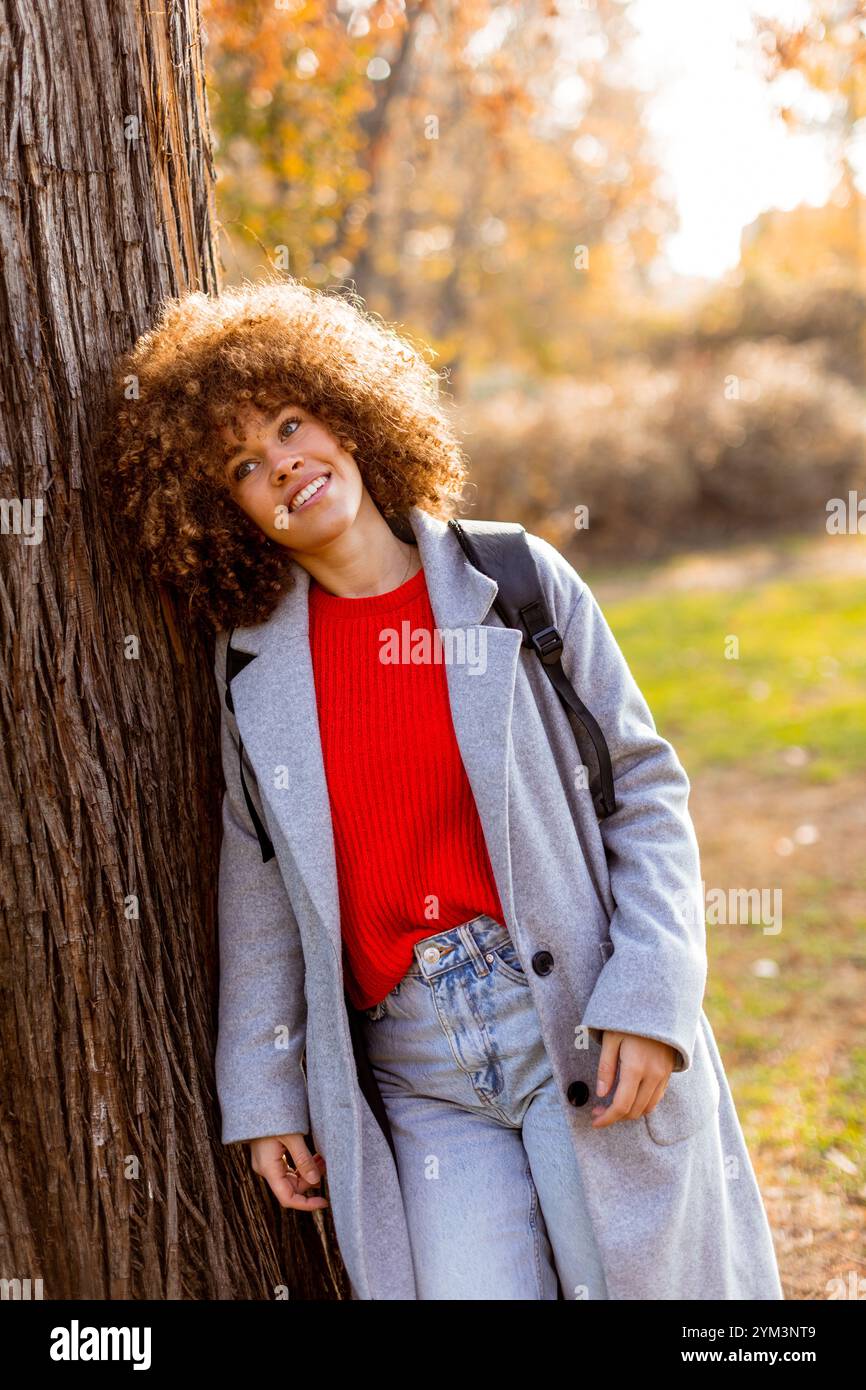 Eine lächelnde junge Frau mit lockigem Haar steht vor einem Baum und trägt ein gemütliches Outfit. Sie liebt die Schönheit des Herbstes in einem ruhigen Park voller Colo Stockfoto