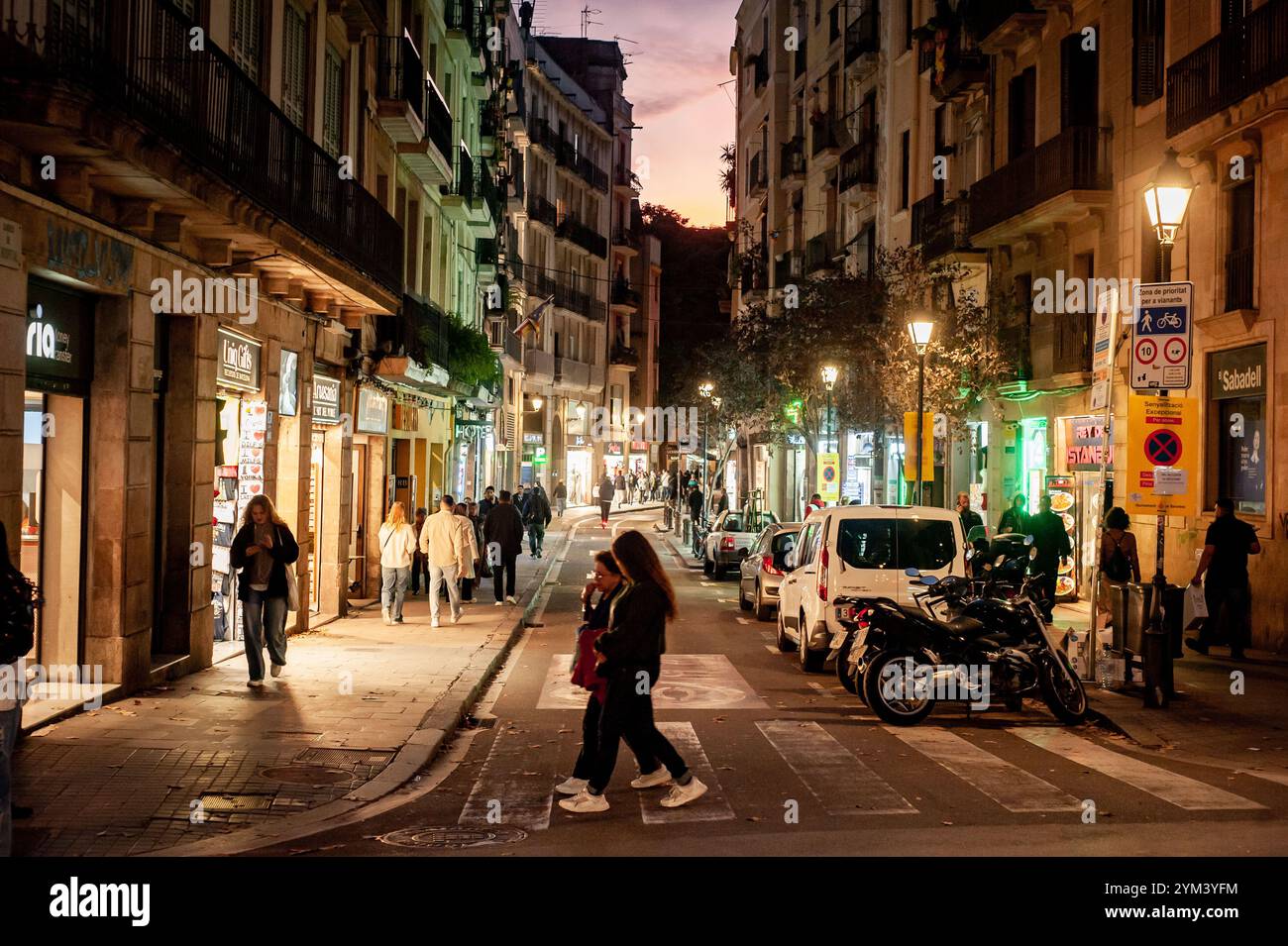 Pendler gehen die Straße des Carrer Hospital neben La Rambla im Viertel Raval entlang, während die Straßenlaternen die Altstadt Barcelonas in der Abenddämmerung erleuchten. Stockfoto