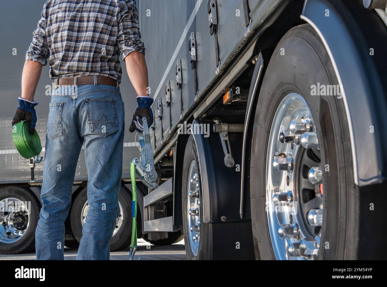 Ein Arbeiter steht in der Nähe eines großen Lkws, hält die Gurte und bereitet sich darauf vor, die Fracht zu sichern, während die Sonne an einer belebten Laderampe untergeht. Stockfoto