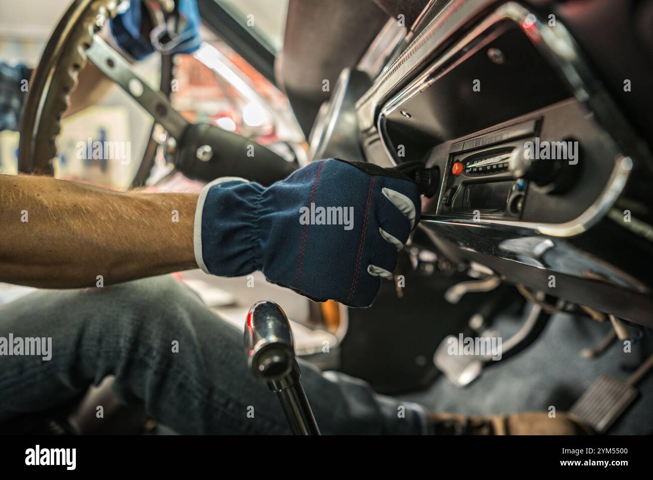 Eine Person passt die Bedienelemente des Armaturenbretts eines Oldtimers an, während sie Handschuhe trägt, umgeben von Werkzeugen und einer Werkbank in einer Garage. Stockfoto