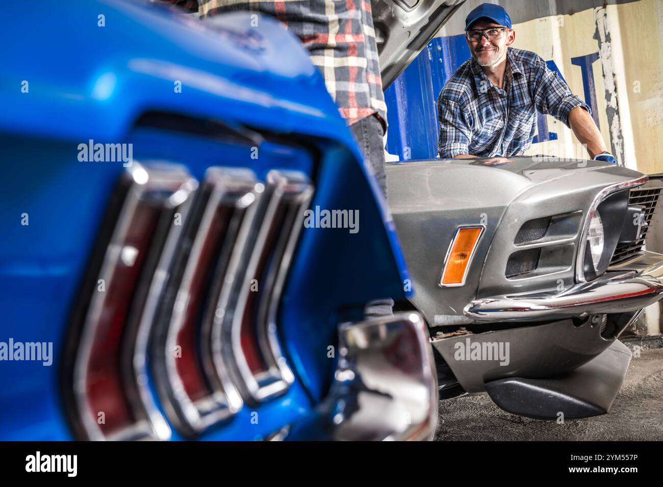 Ein Mechaniker arbeitet an einem Oldtimer in einer gut beleuchteten Garage und konzentriert sich dabei auf Motorreparaturen, während ein anderes Auto im Vordergrund sichtbar ist. Stockfoto