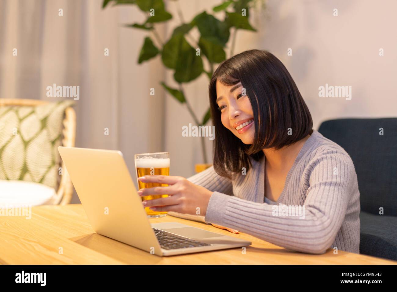 Eine junge Frau trinkt Bier, während sie nachts im Zimmer auf den Computer schaut. Stockfoto