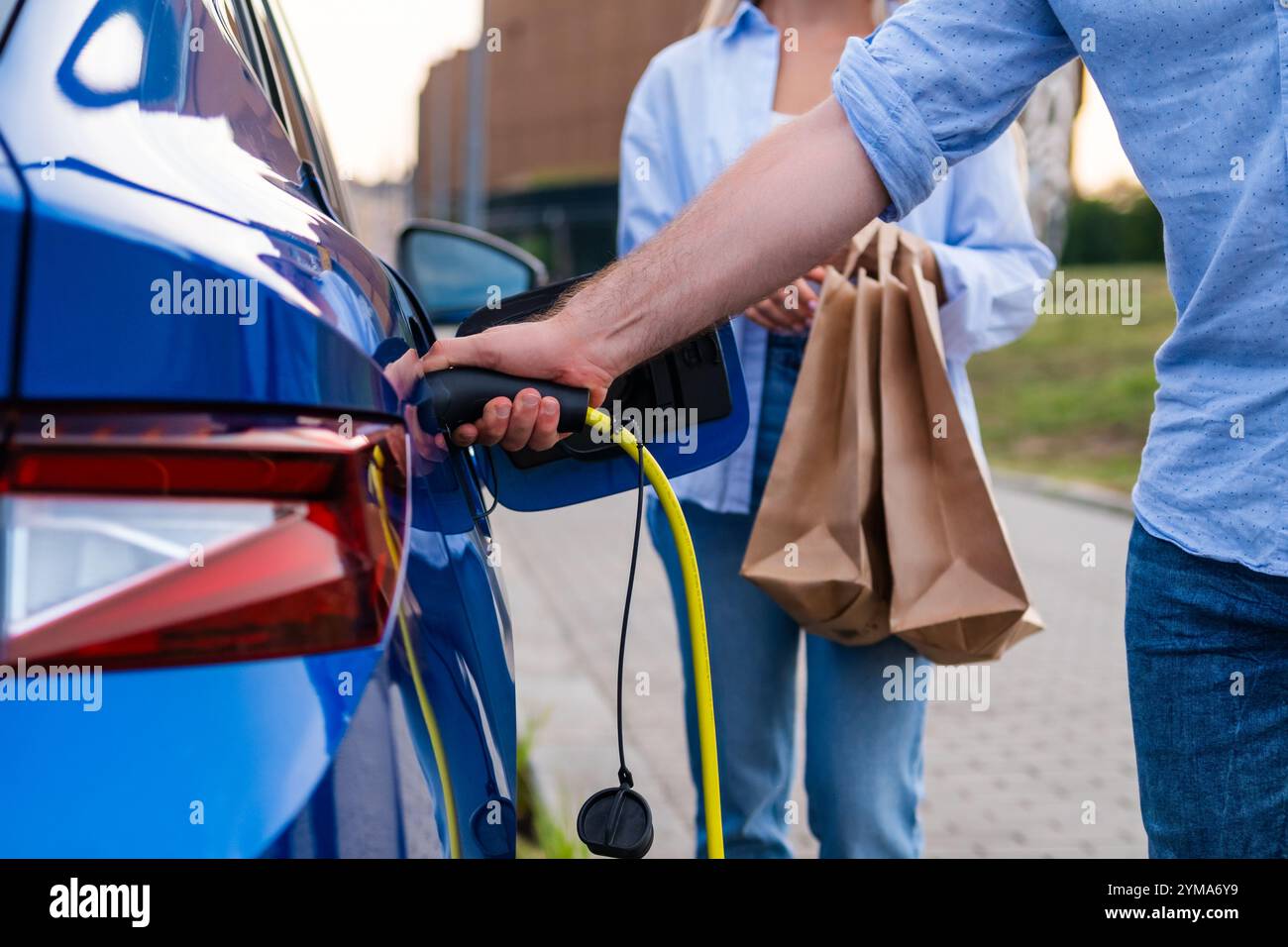 Eine Person lädt ein Elektroauto auf, während ein anderer auf einem städtischen Parkplatz Einkaufstaschen hält Stockfoto