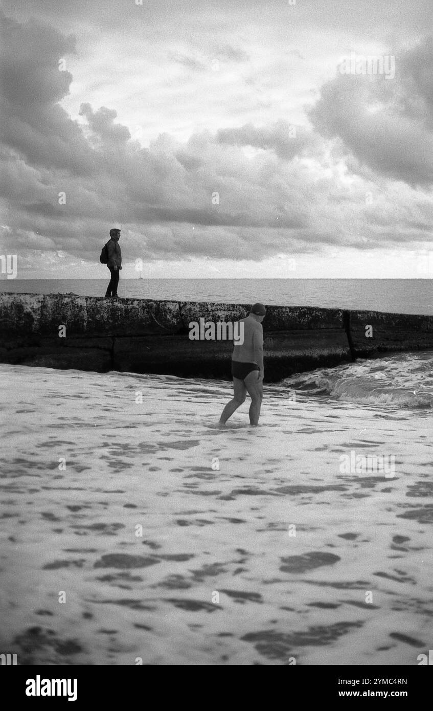 Ein dramatisches Schwarzweiß-Foto, das eine Küstenszene mit einer Person darstellt, die im Wasser steht und eine andere auf einem Betonpier, auf einem Rücken Stockfoto