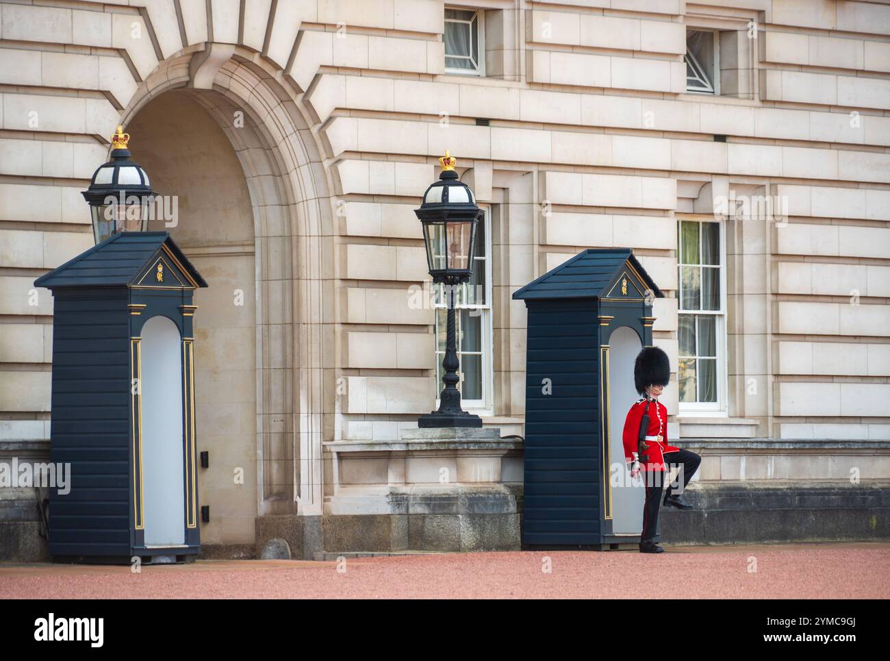 Ein Zeremoniellwächter in einer roten Tunika und einem Bärenfellhut steht Wache und marschiert in der Nähe von Wachhäusern vor dem Buckingham Palace in London. Stockfoto
