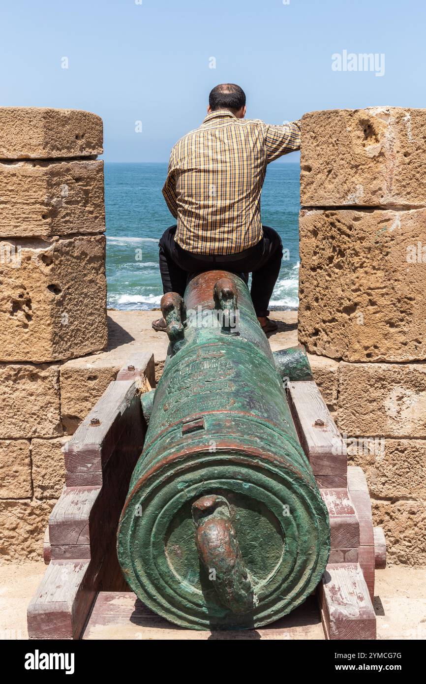 Auf einer alten Kanone in der Festung Essaouira steht ein Mann, der das Meer betrachtet Stockfoto