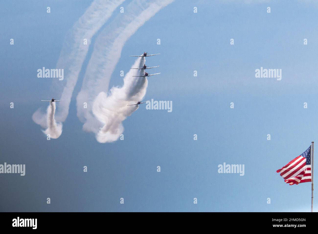Fünf Flugzeuge erzeugen Rauchspuren, während sie in Formation neben einer schwenkenden amerikanischen Flagge vor einem blauen Himmel fliegen. Stockfoto