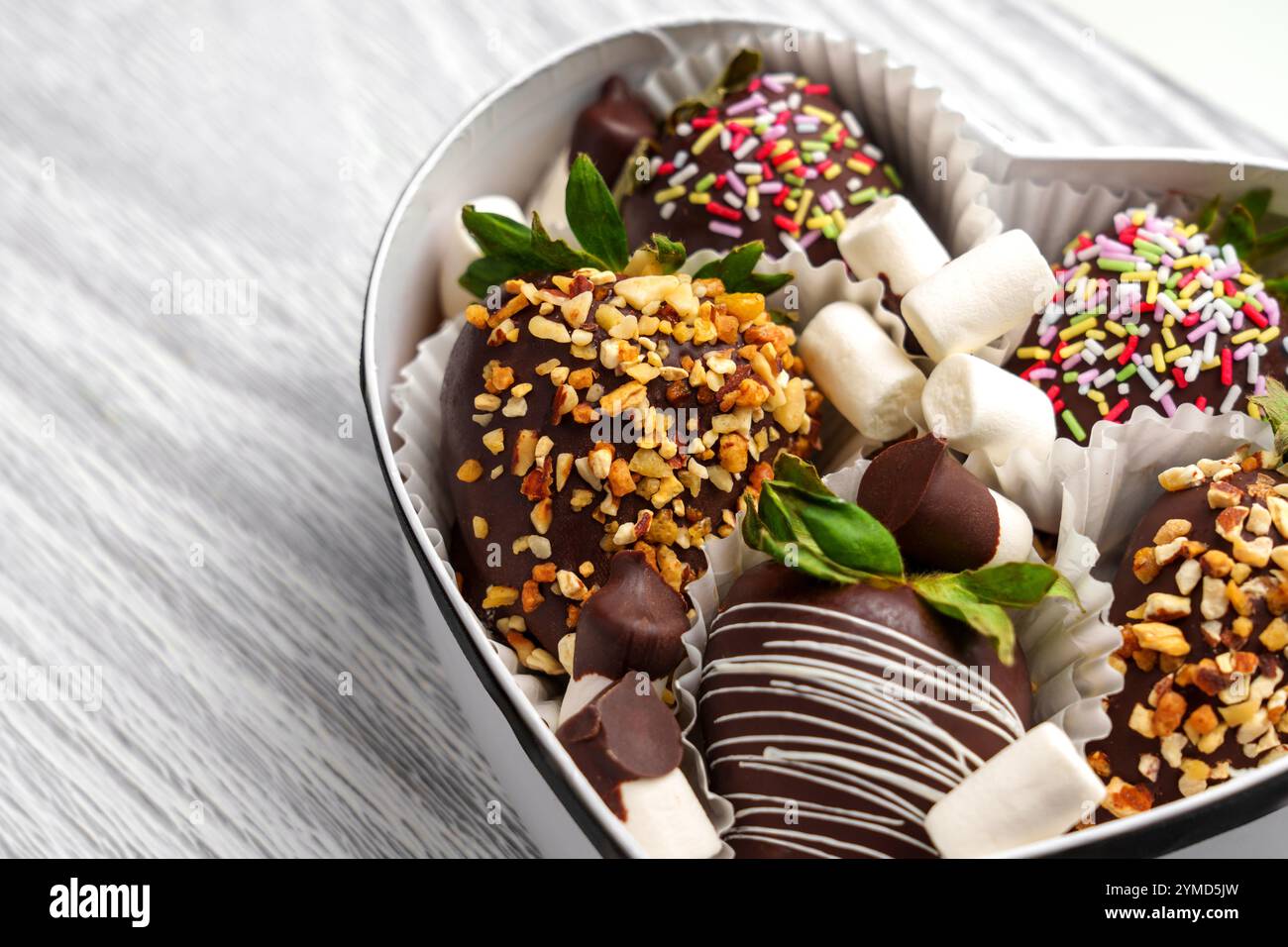 Handgefertigtes Erdbeerdessert, beschichtet mit zerkleinerten Nüssen und weißer Glasur im Papierbehälter. Frische grüne Blätter sorgen für einen lebendigen Touch. Für festlichen Look Stockfoto