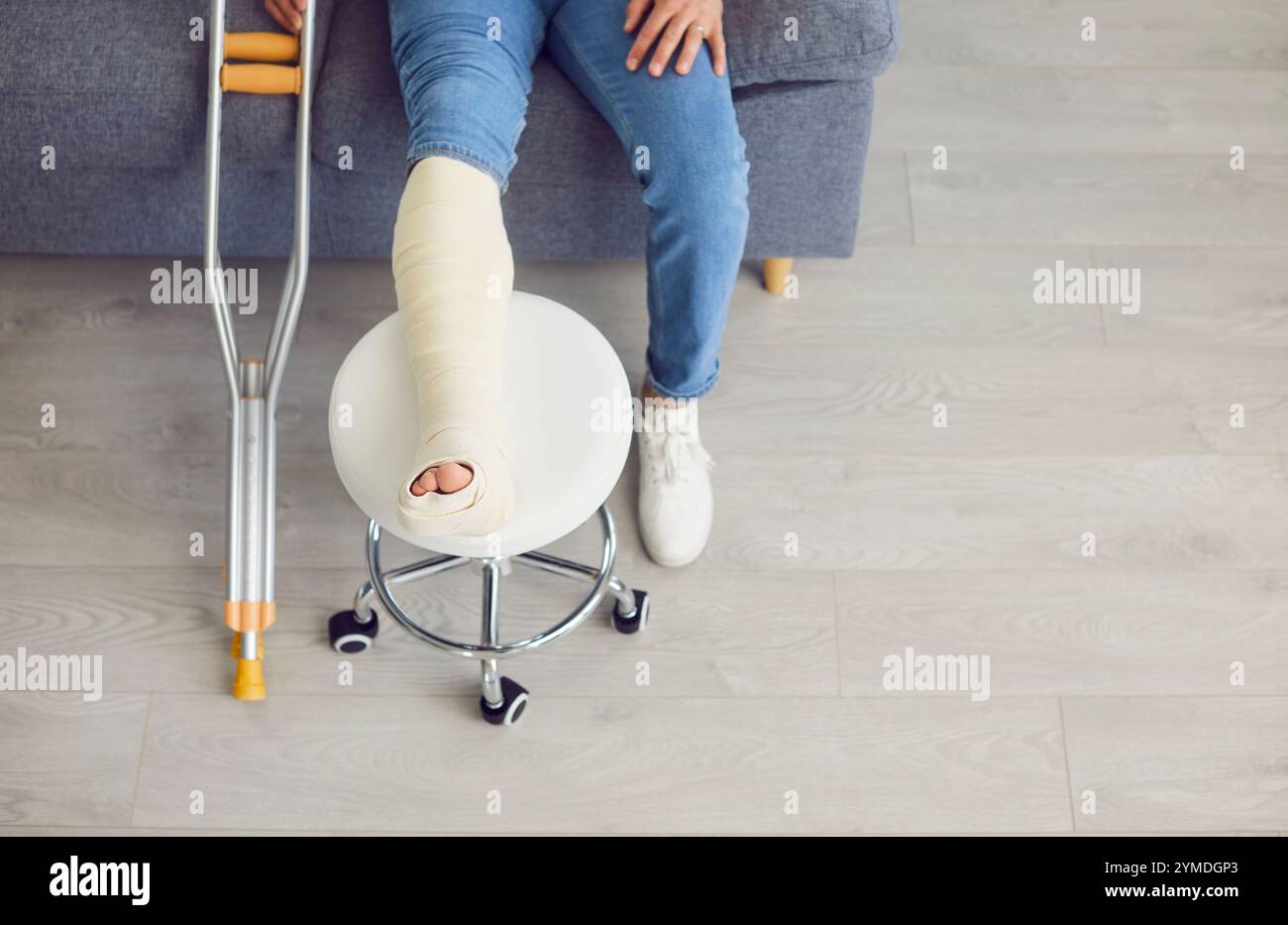 Nahaufnahme von gekrümmten Krücken und gebrochenem, bandagiertem Bein aus Gips auf dem Stuhl. Draufsicht Stockfoto